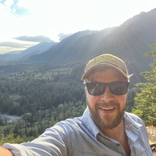 A selfie of Ben on a sunny hike wearing a button-up shirt, sunglasses, and ball cap.