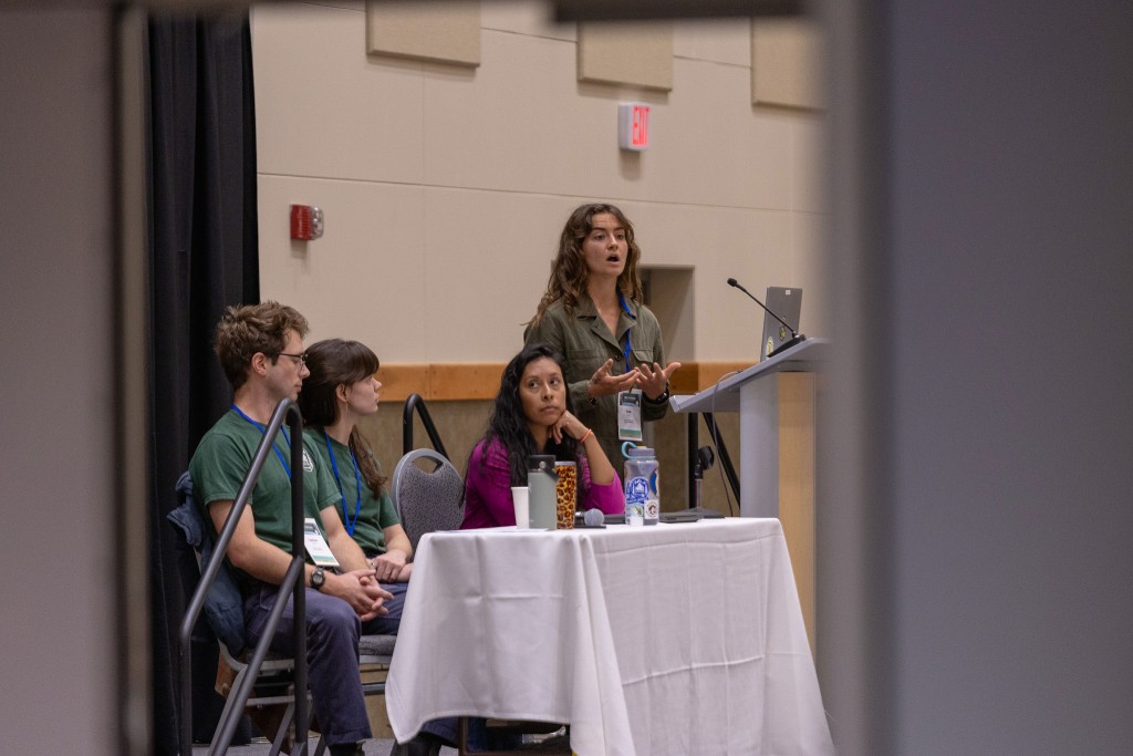 A panel of presenters stands at a raised table while speaking to an audience.